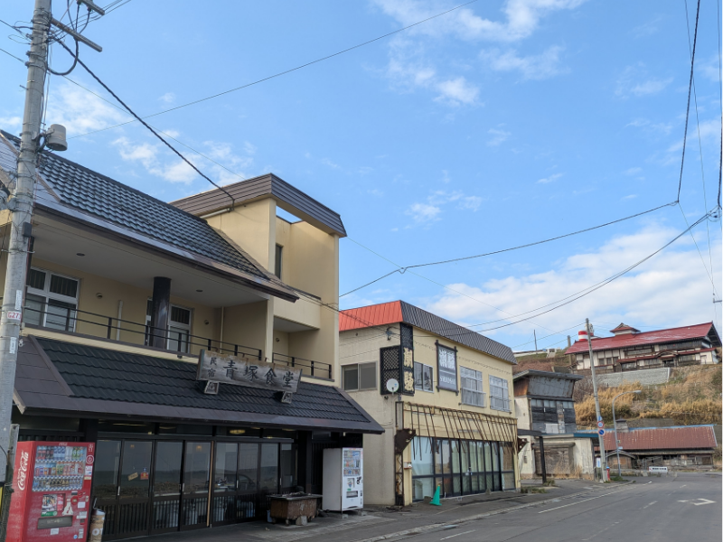 小樽・祝津にある「民宿 青塚食堂」の店舗外観。青空の下、看板メニューであるニシンの炭火焼きの香りが漂ってきそうな、歴史を感じる建物の様子。