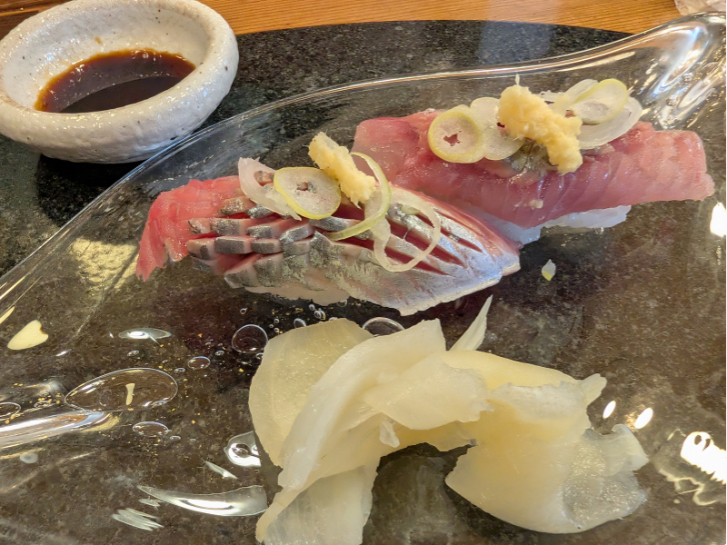 透明な器に盛り付けられたニシンの握り寿司2貫。飾り包丁が入った美しい身に、生姜とネギが添えられ、手前にはたっぷりのガリが添えられている様子。