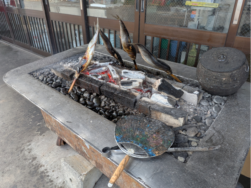 「民宿 青塚食堂」の店先で、大きなニシンを串に刺し、炭火の周りに立てて豪快に焼き上げる様子。使い込まれた団扇（うちわ）が手前に置かれている。