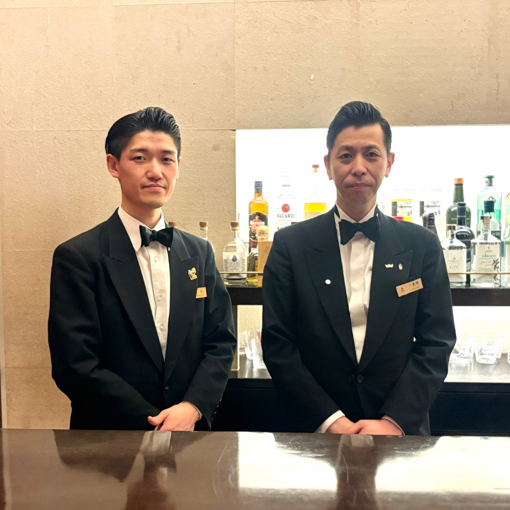 カウンターの中で並んで立つ、正装した二人のプロフェッショナルなバーテンダー