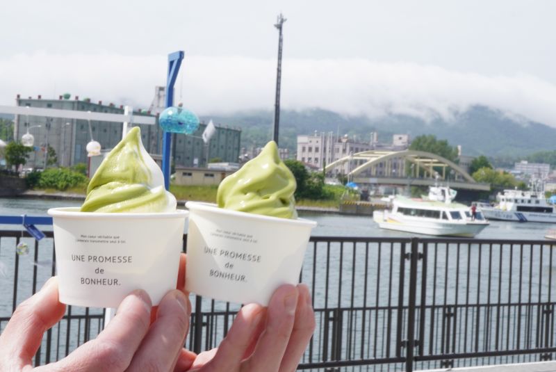 小樽の海と港の景色をバックに、手で持った「Port bakery nomnom」の宇治抹茶ソフトとミックスソフトのカップが2つ並んでいる写真