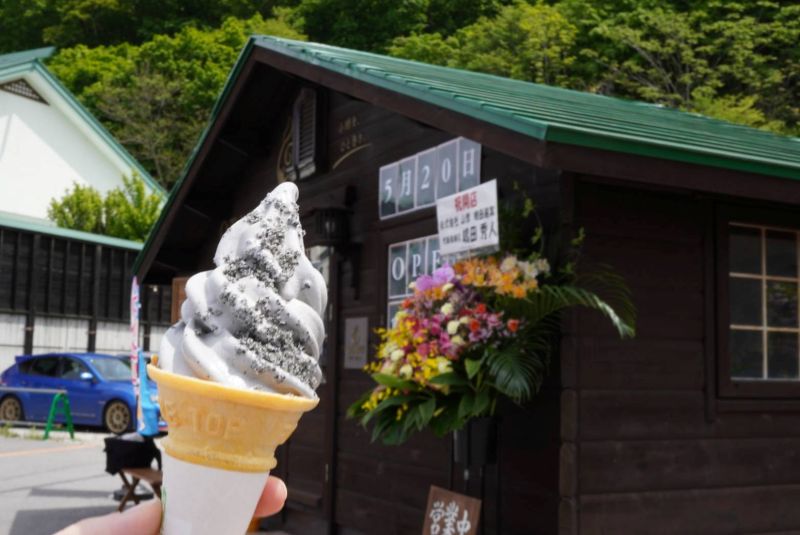 ログハウス風の建物をバックに、黒いごまがたっぷりとトッピングされた「OTARU SOFT CREAM」のグレー色の朝里ダムソフトを手に持っている写真