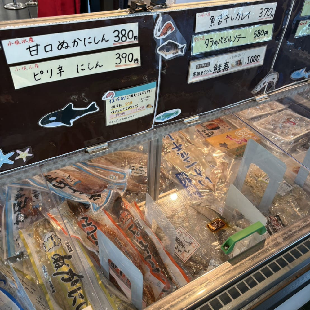 道の駅みなとま～れ寿都内の鮮魚コーナーにある冷凍ショーケース。ケース上部には「甘口ぬかにしん」「タラのバジルソテー」「鮭寿」といった寿都産の加工品を紹介する手書きの値札ポップが掲示されており、ケース内にはパックに詰められた様々な鮮魚や加工品が並んでいる様子。