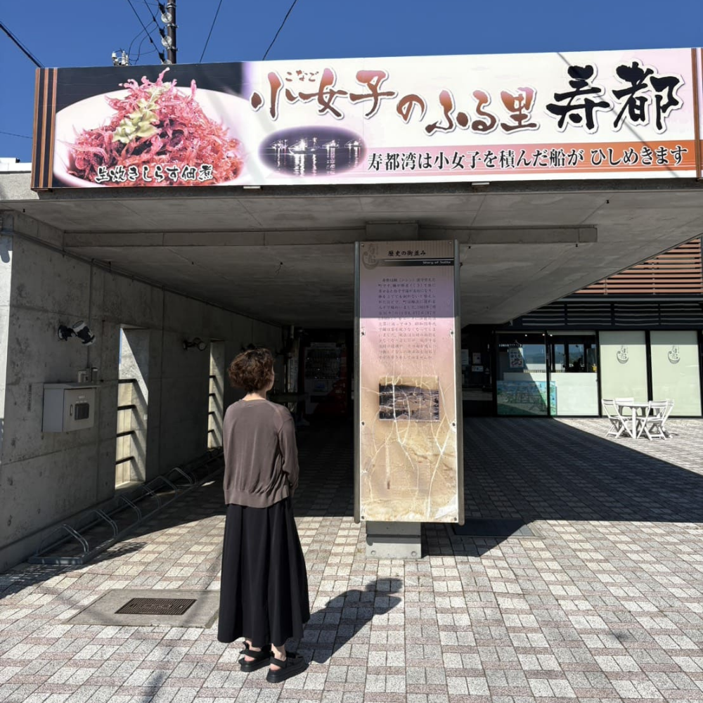 寿都町にある「道の駅みなとま～れ寿都」の入口。上部には「小女子のふる里 寿都」と書かれた、生炊きしらす佃煮や漁船の様子が描かれた大きな看板があり、その下で女性が立ち止まって案内板を眺めている様子。