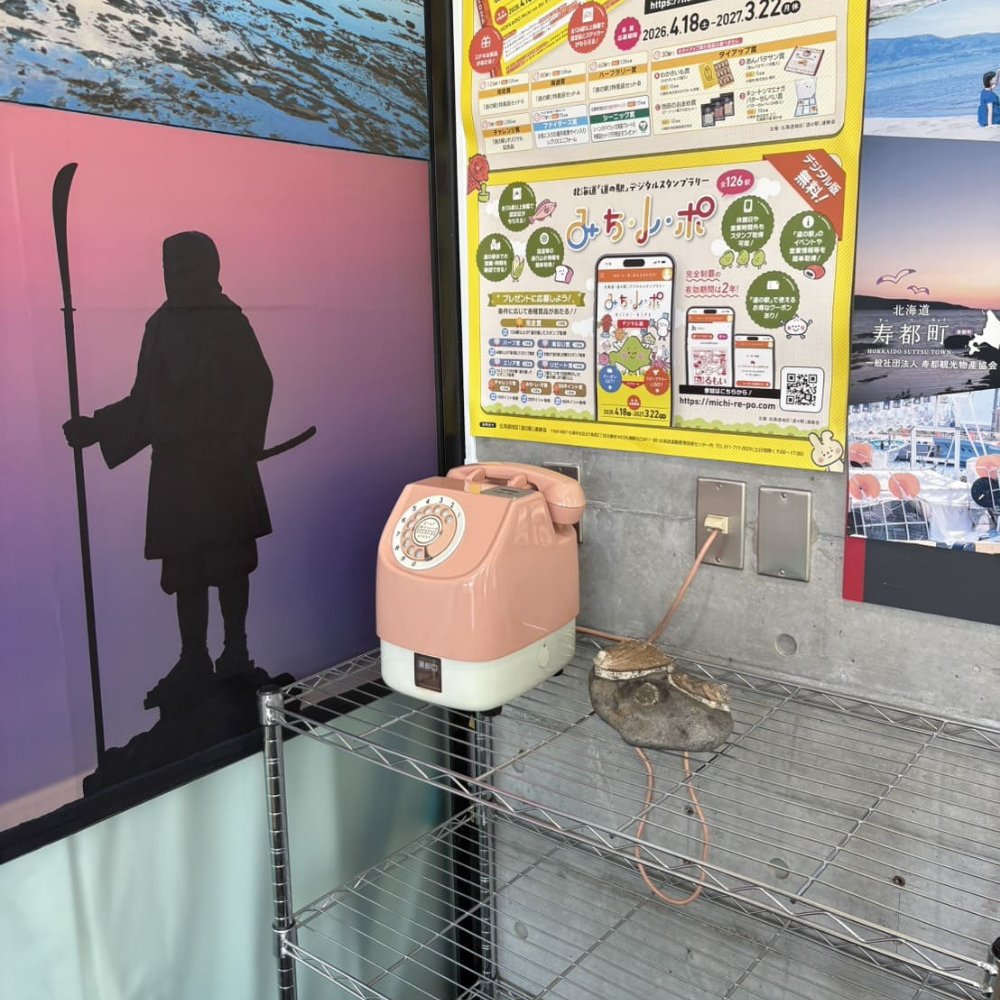 道の駅みなとま～れ寿都内の一角。武蔵坊弁慶のシルエットが描かれた紫色の夕日を背にするポスターと、隣には寿都町やデジタルスタンプラリー「みち・レ・ポ」のポスターが掲示されている。手前のワイヤーラックには、レトロなピンク色のダイヤル式電話機と、その横に置かれた貝殻が写っている様子。