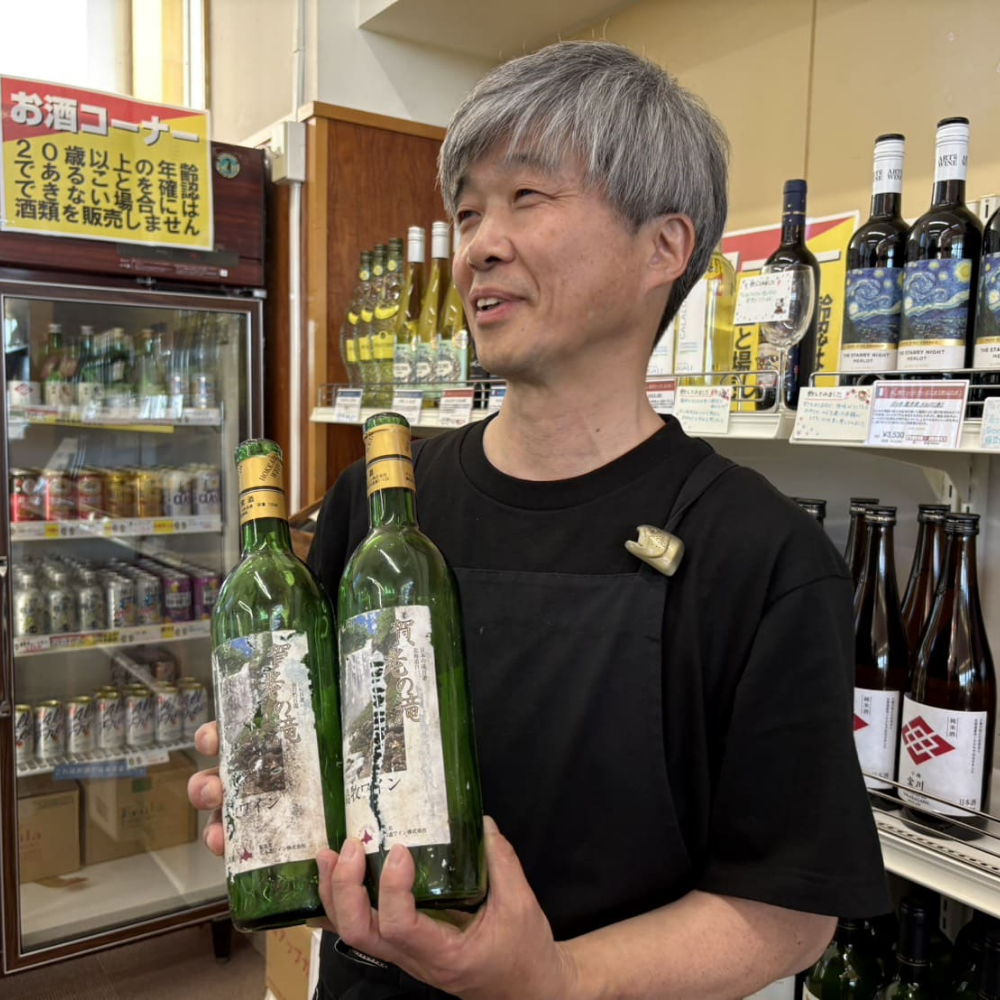 店内の「お酒コーナー」にて、過去に製造された「賀老の滝」ラベルの島牧ワイン（緑色のボトル）を両手に持ち、微笑む男性（店主または地域おこし協力隊員）。背景には地酒やワインが整然と並ぶ棚が写っている様子。