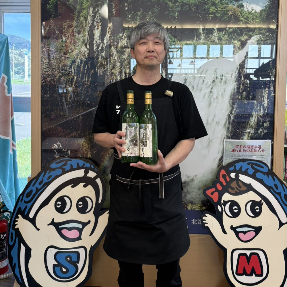 旧「島牧ワイン」のボトル2本を持ち、笑顔で立つ男性。背景には大きな「賀老の滝」の写真パネルが掲げられ、手前両脇には島牧村のシンボルキャラクター（島牧村のキャラクター看板）が配置されている様子。