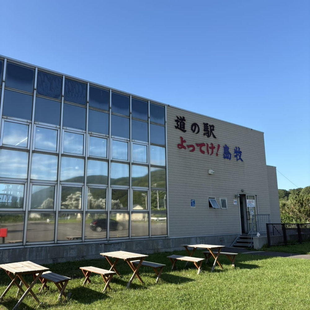 青空の下に立つ「道の駅 よってけ！島牧」の建物外観。大きなガラス張りの壁面と、壁に書かれた施設名の文字、手前には木製のベンチやテーブルが設置されている様子。