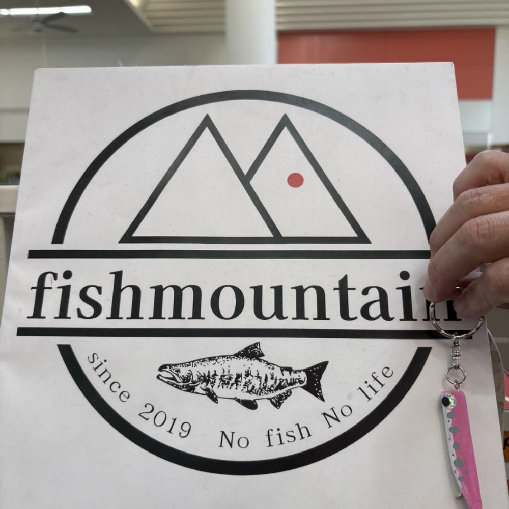 「fishmountain」のロゴが印刷された白い紙袋。山をモチーフにしたマークと「No fish No life」「since 2019」の文字、魚のイラストが描かれており、右端には手で掲げられた魚の形をしたピンク色のルアーキーホルダーが写っている様子。