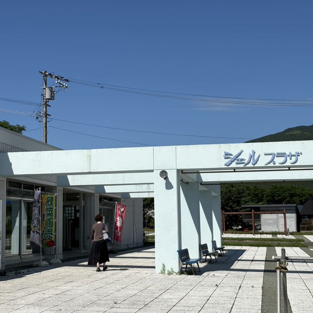 寿都町にある物産館「シェルプラザ・港」の外観。青い文字の施設名サインと、手前に立つ「野菜直売」ののぼり、建物へ向かう女性の後姿
