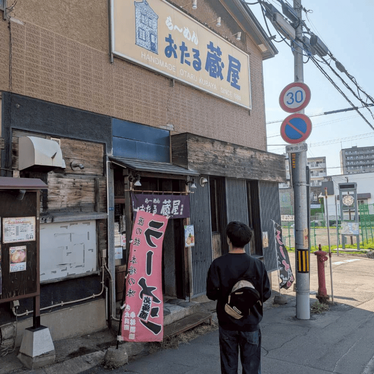 「ら〜めん おたる蔵屋」の店舗外観と、お店に向かうリュックを背負った男性の後ろ姿