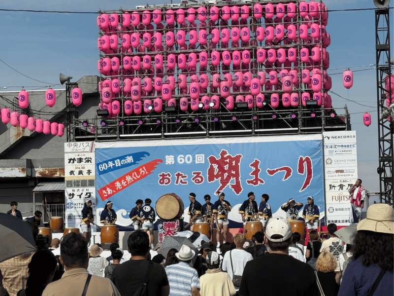 「第60回おたる潮まつり」の看板と多数のピンクの提灯が飾られた中央ステージで、潮太鼓の打ち手たちが演奏を披露する様子