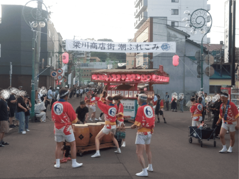 梁川商店街の入り口に掲げられた「梁川商店街 潮ふれこみ」の横断幕の下で、赤い法被を着た打ち手たちが山車の前で潮太鼓を力強く打演する様子