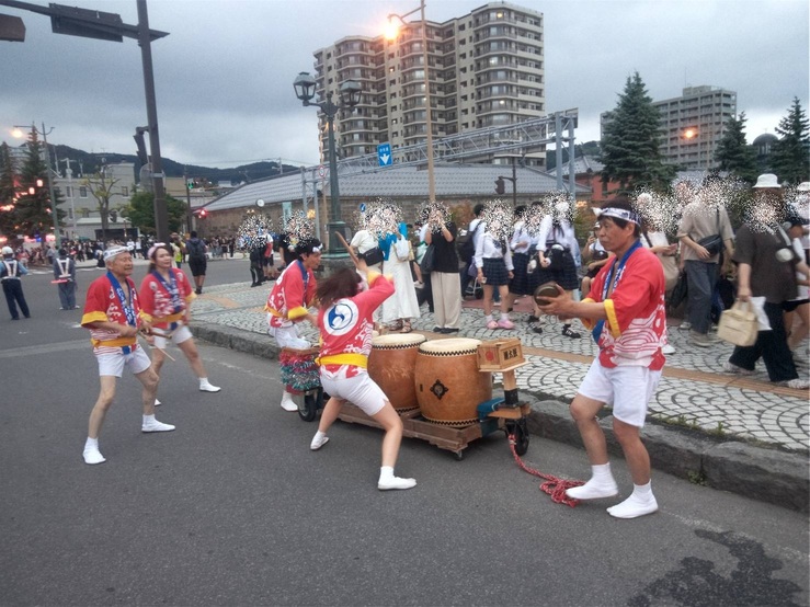 石造り倉庫群が広がる小樽運河浅草橋付近の路上で、赤い法被を着た保存会の人々が台車に乗せた太鼓や鉦(かね)を威勢よく演奏する様子