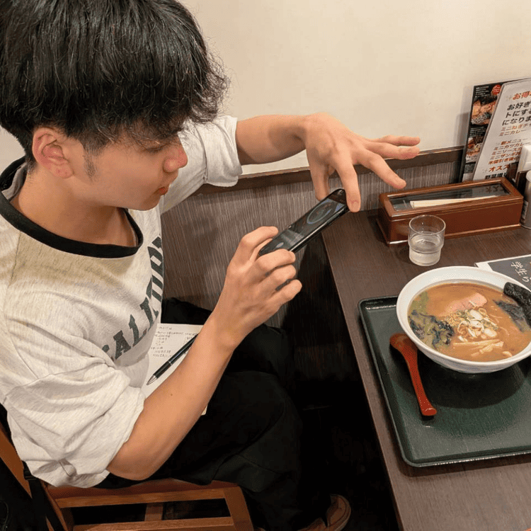 「らーめん むくげ」の店内で、提供されたラーメンをスマートフォンで熱心に撮影する大学生