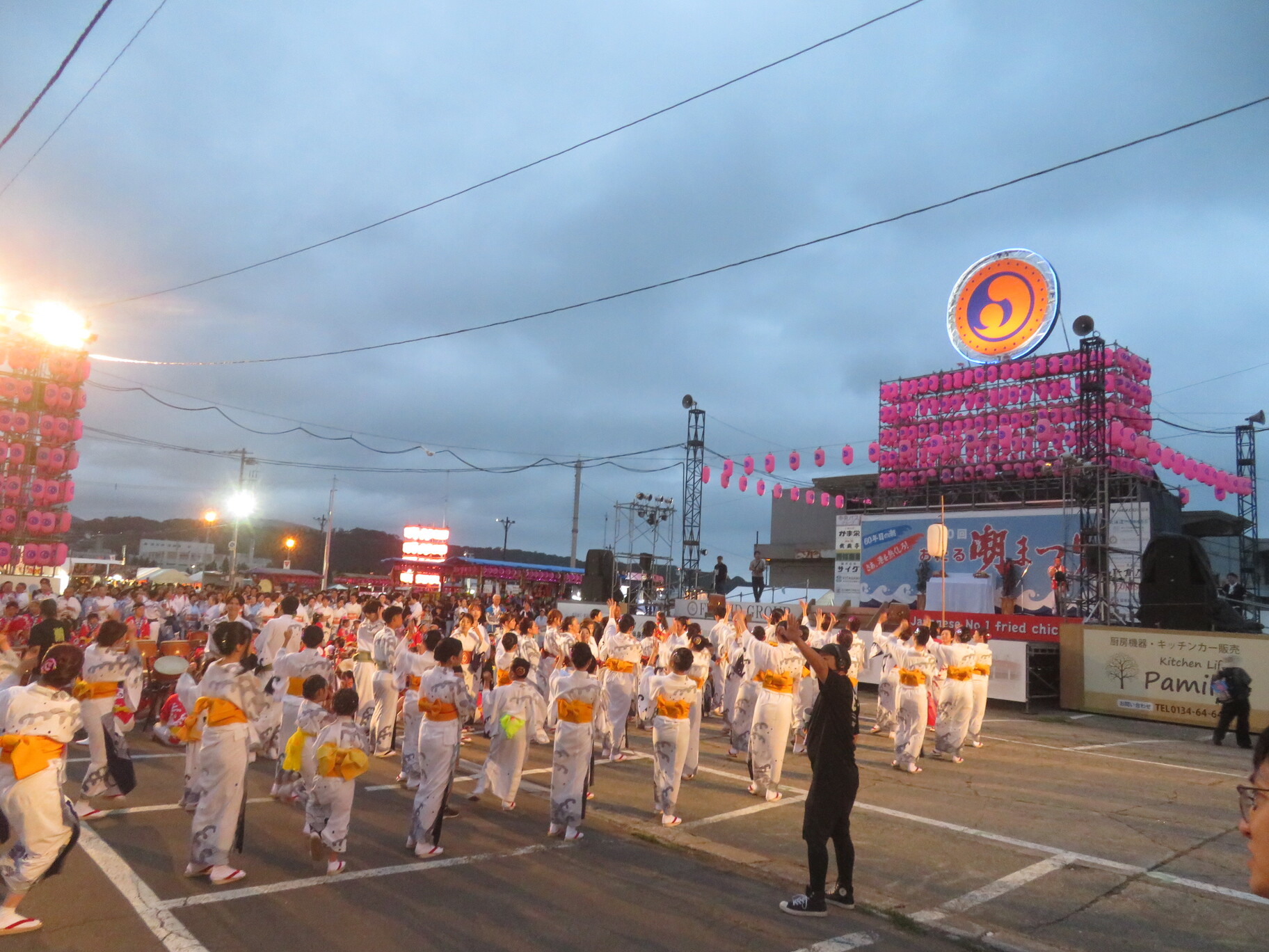 第60回おたる潮まつりのメインステージ前で、ライトアップされた提灯や看板を背景に、揃いの浴衣を着た大勢の参加者が輪になって潮音頭を踊る様子