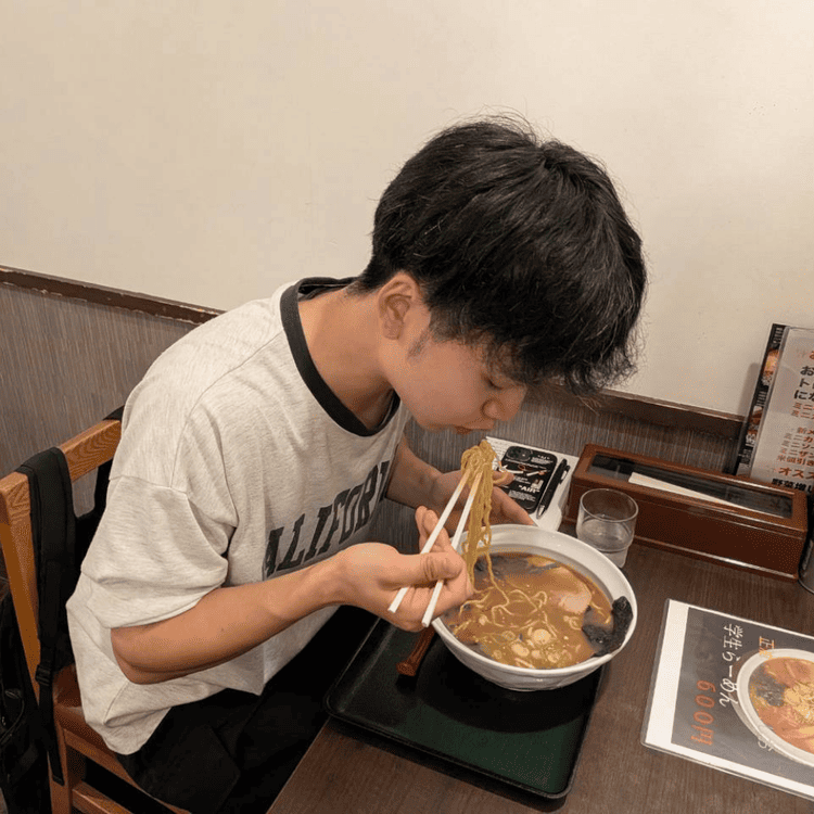 「らーめん むくげ」の店内で、箸で持ち上げたラーメンの麺を勢いよくすする大学生