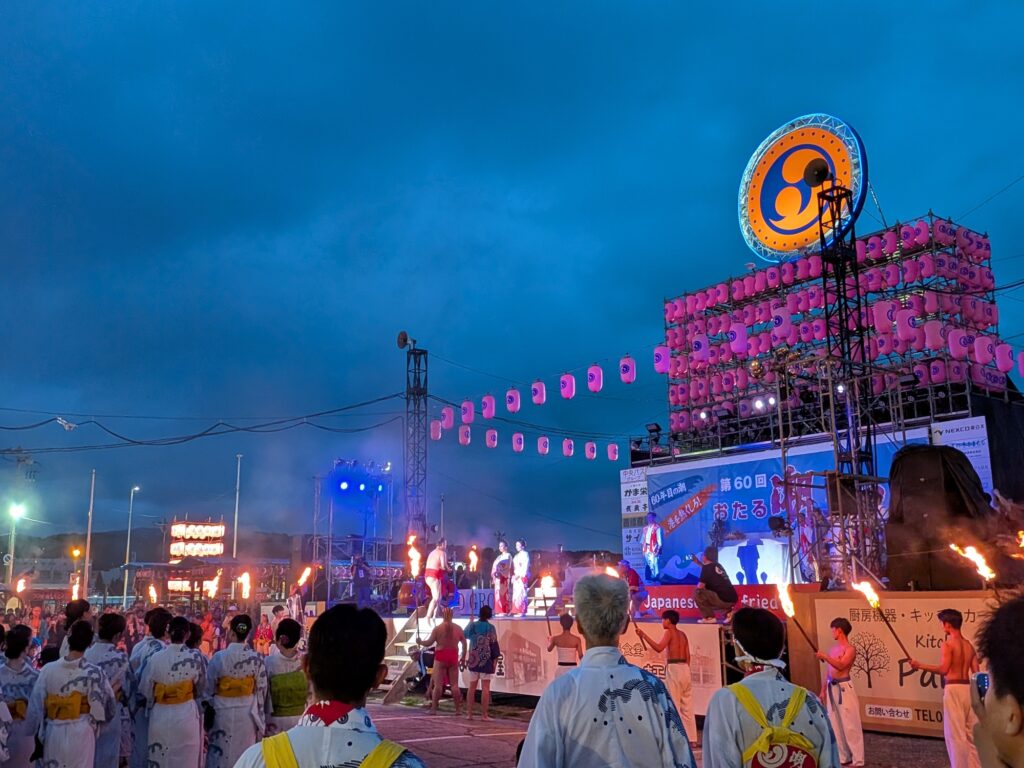 夜のおたる潮まつりメインステージ前で、松明を持った男性たちや浴衣姿の参加者が見守る中、ステージ上で御神火が奉納される様子