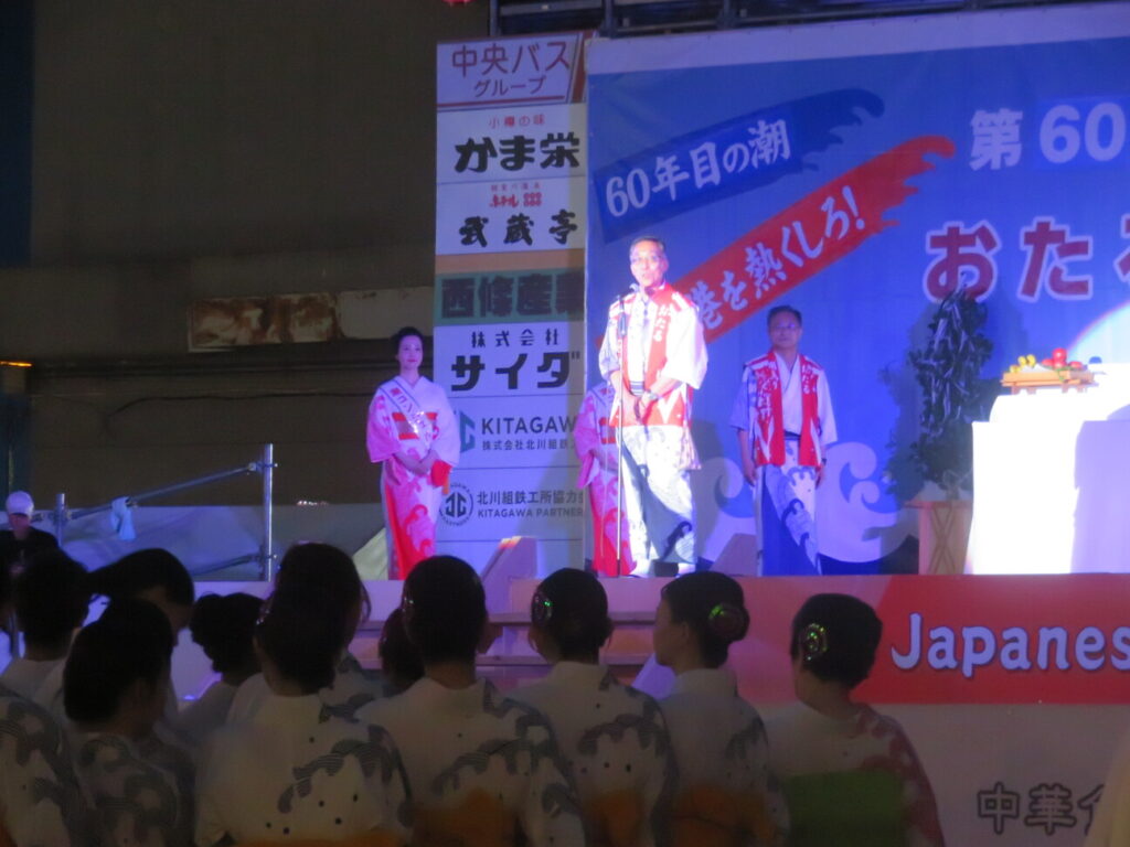 第60回おたる潮まつりのメインステージ上で、浴衣姿の参加者や潮コンシェルジュが見守る中、マイクの前に立ち開会の挨拶を行う迫市長の様子
