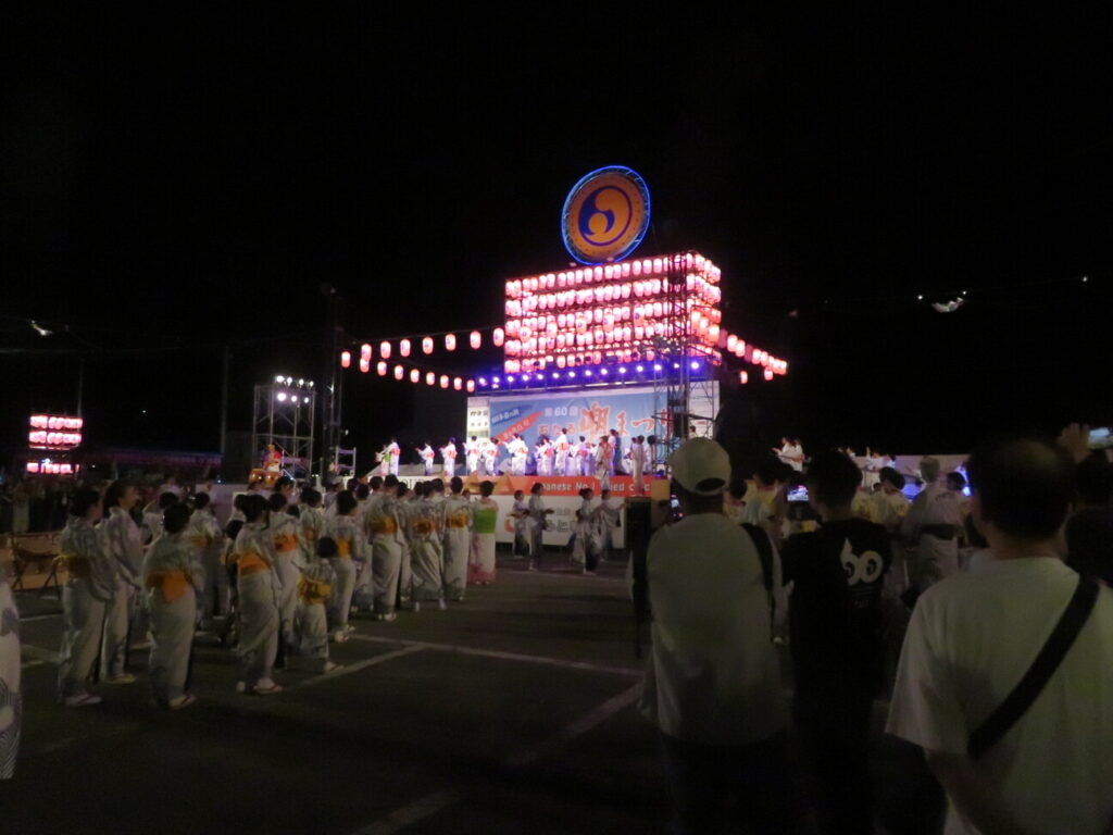 無数の提灯とシンボルマークが鮮やかに輝く夜のおたる潮まつりメインステージ前で、浴衣姿の参加者たちが潮音頭を踊り、手前で大勢の観客が見守る様子