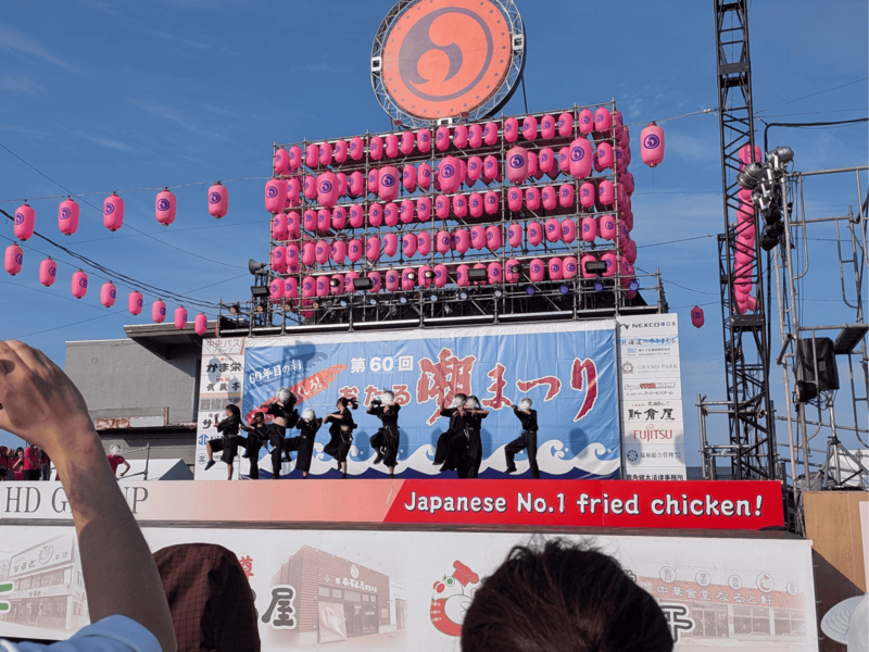 「第60回おたる潮まつり」の巨大看板とピンクの提灯が飾られた野外ステージで、黒い衣装を着たダンサーたちがダンスパフォーマンスを行っている様子
