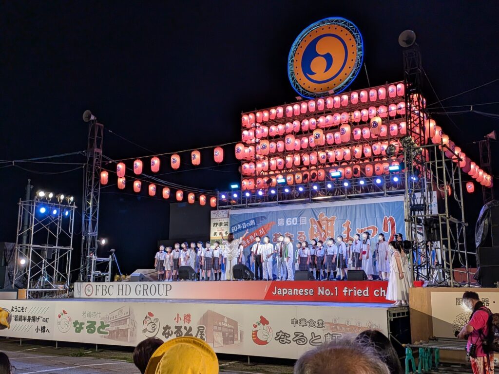 無数の提灯とマークが輝く夜の「第60回おたる潮まつり」メインステージ上で、制服や衣装を着た子どもたちが並んで合唱を披露する様子