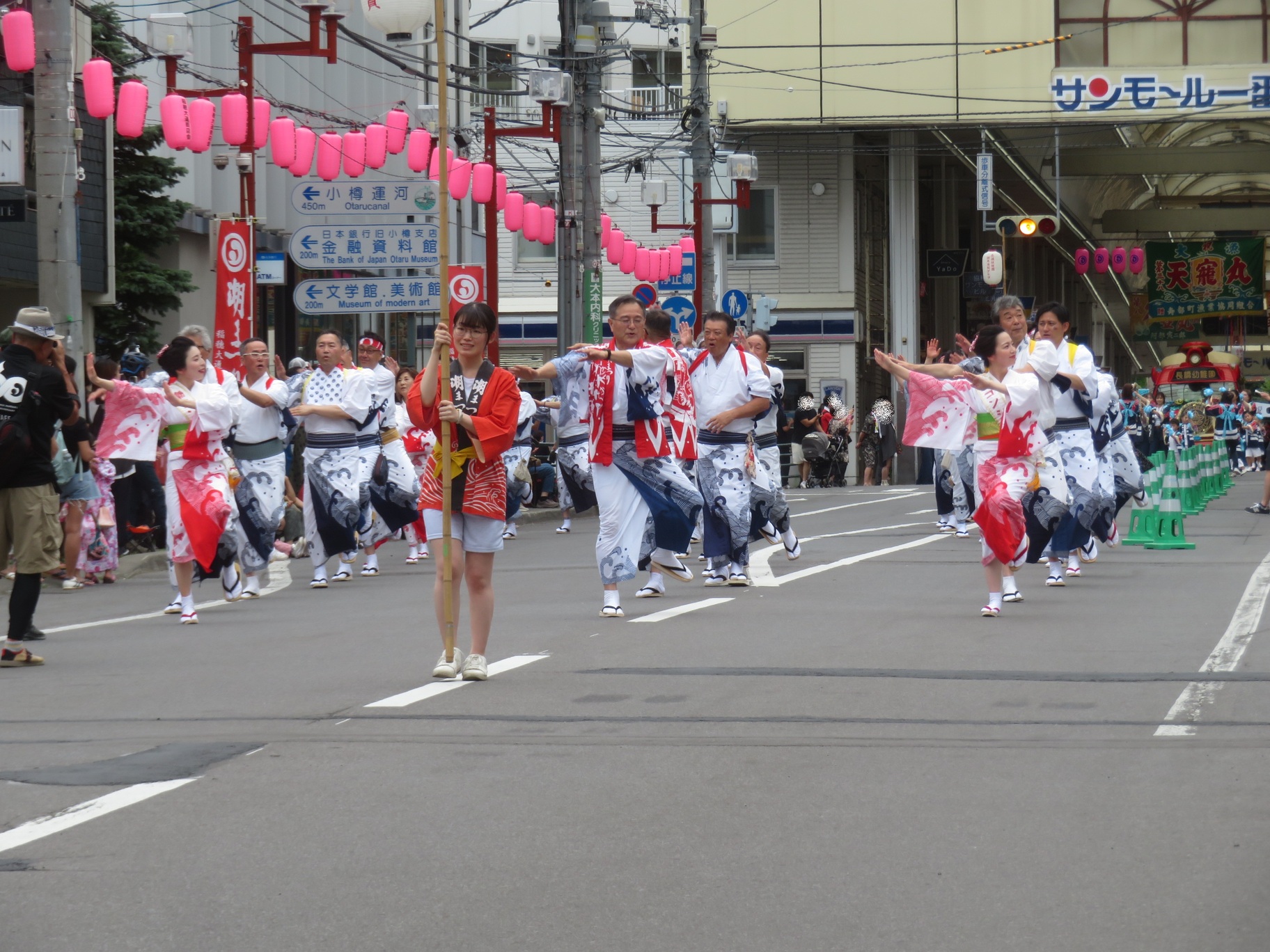サンモール一番街のアーケード入口付近の道路で、長い竹竿を持った女性を先頭に、潮コンシェルジュや関係者、浴衣姿の参加者たちが潮音頭を踊りながら進む様子