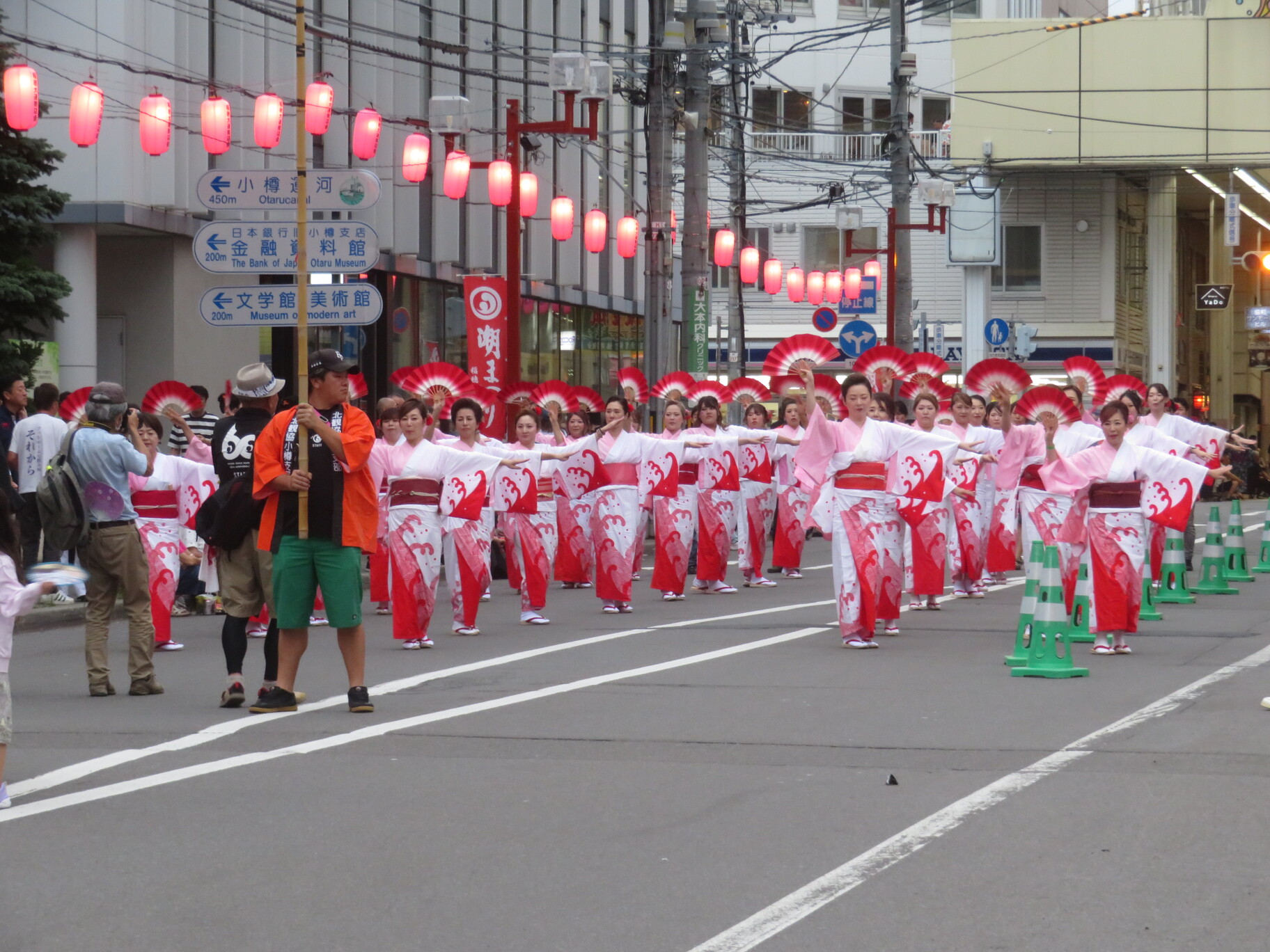 赤い提灯が吊るされた街路で、白と赤の波模様の浴衣を着た女性たちが、鮮やかな赤い扇子を手に揃った手振りで踊り進む潮ふれこみの様子