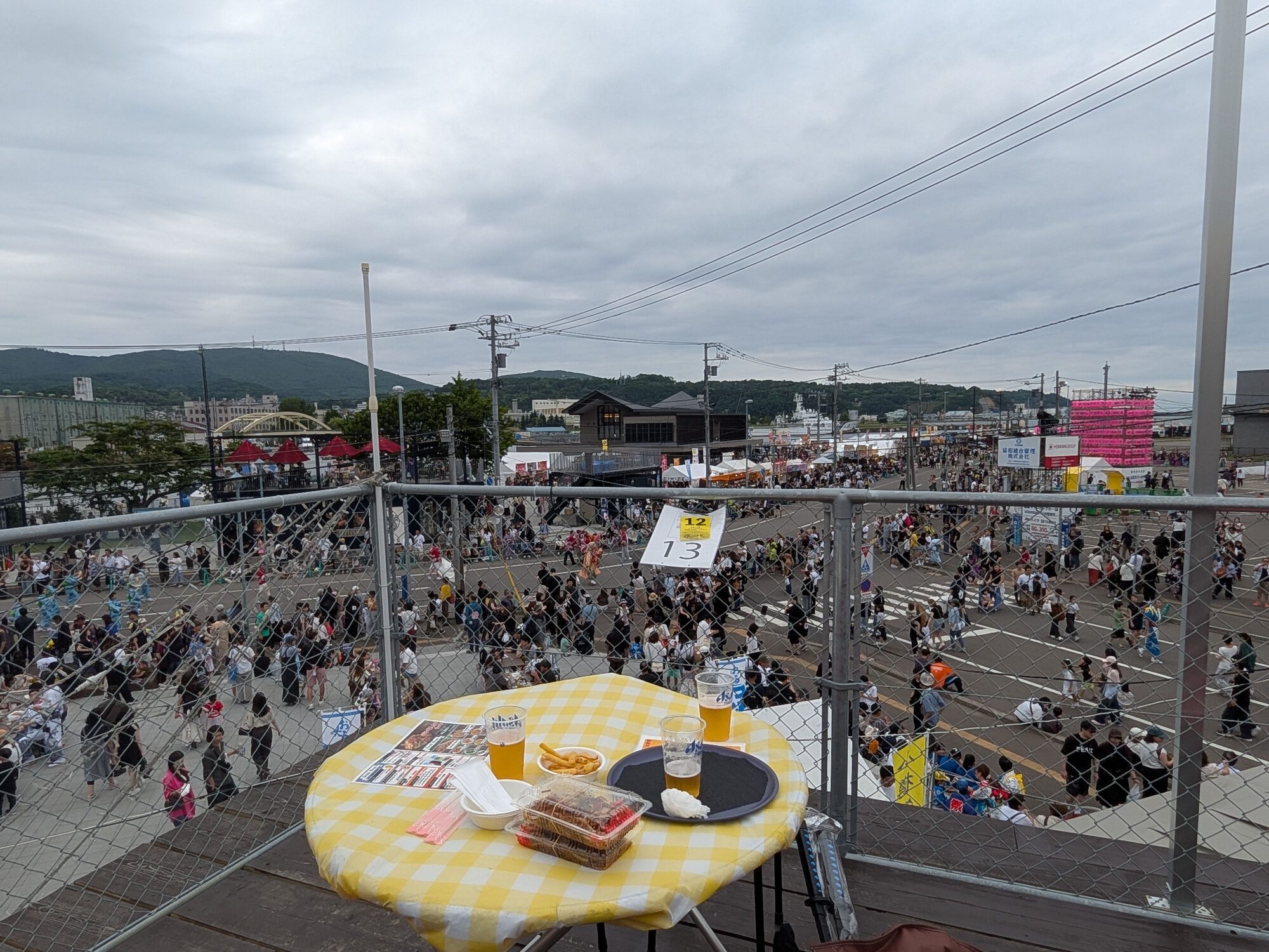 黄色と白のチェック柄のクロスが敷かれたハイボールテラスの丸テーブル越しの眺めで、ドリンクや軽食の向こうに大勢の来場者で賑わうおたる潮まつり会場全体を見渡す様子