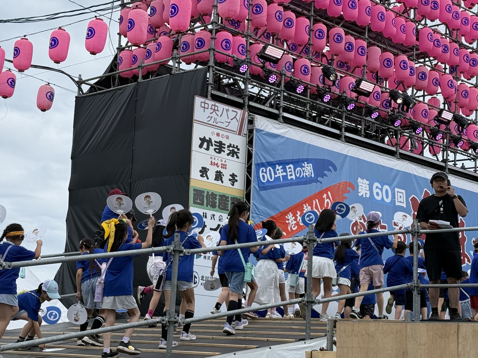 ピンクの提灯が飾り付けられた「第60回おたる潮まつり」のメインステージのスロープを、揃いの青いTシャツを着てうちわを手にした大勢の子どもたちが登っていく様子