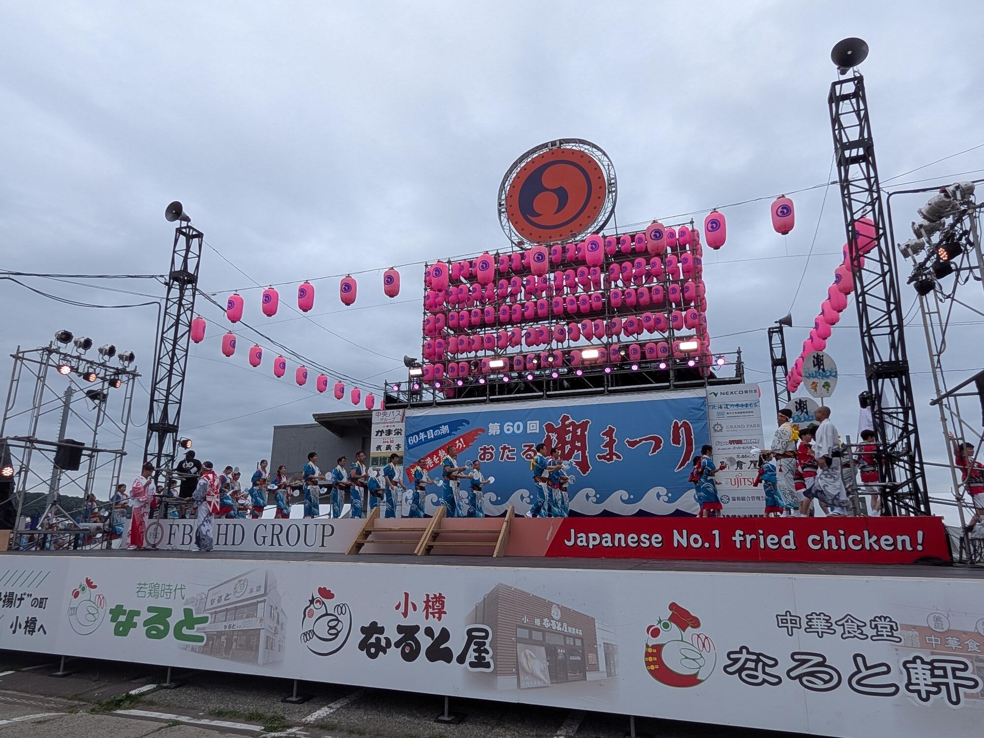 日中の「第60回おたる潮まつり」メインステージ全景。提灯や大きなシンボルマークが飾られたステージ上で、青い踊り衣装を着た出演者たちが一列になって演舞を披露する様子