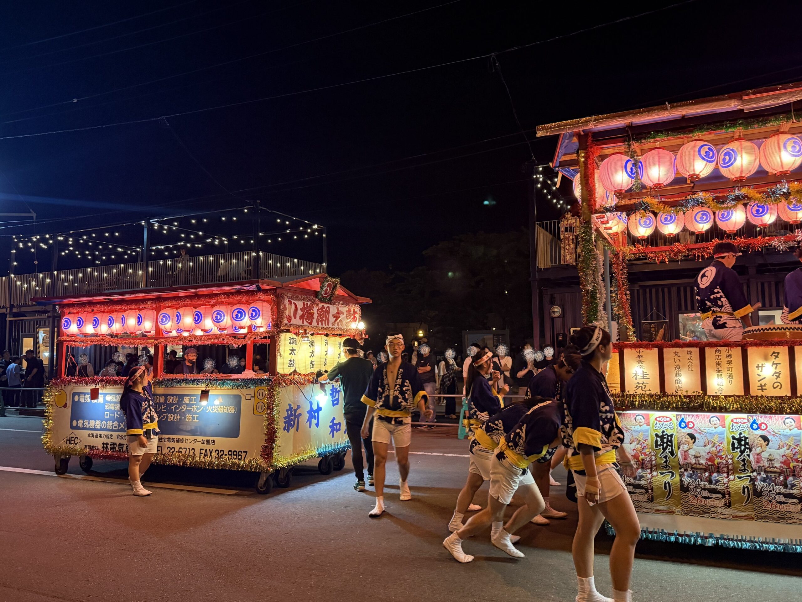 夜間のおたる潮まつり会場で、赤や白の提灯や電飾で華やかに彩られた2台の大きな山車の周囲を、紺色の半纏と白い半股引きを着た小樽潮太鼓保存会のメンバーたちが引いて進む様子