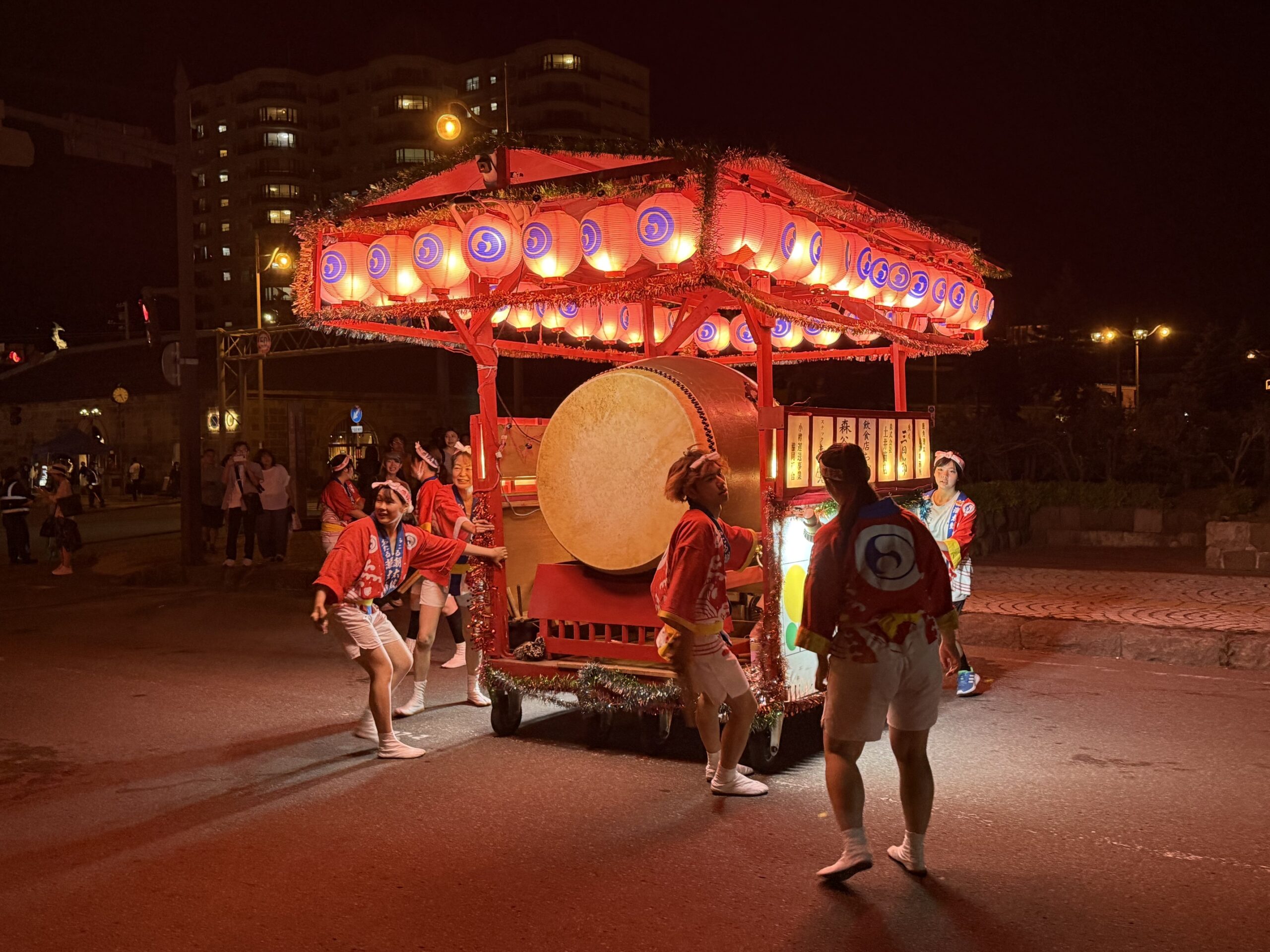 夜間の路上で、あたたかみのある赤い提灯がズラリと点灯した大太鼓付きの山車を、赤い法被を着た保存会の若者たちが取り囲み、腰を落として力強く引き進む様子