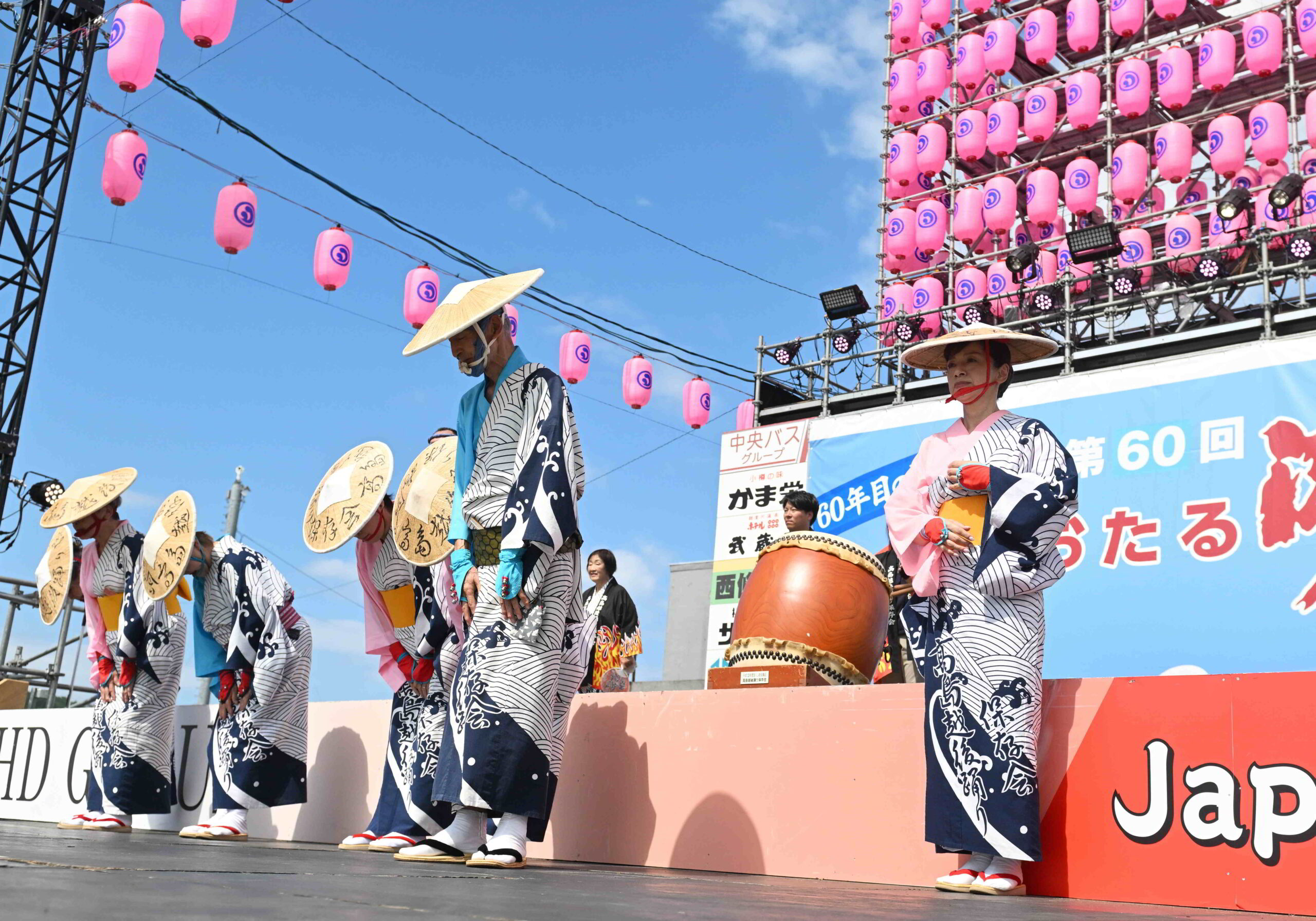 青空とピンクの提灯が広がるおたる潮まつりのメインステージ上で、「高島越後盆踊り保存会」と書かれた波模様の浴衣に編み笠を身にまとった踊り手たちが、演技前後に一斉にお辞儀をする様子