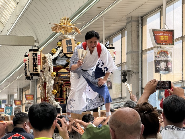 サンモール一番街のアーケード内で、「朝里神社」の提灯が下がった神輿の上に乗り、白い着物姿で扇子を手にして威勢よく群衆を盛り上げる男性と熱気あふれる渡御の様子