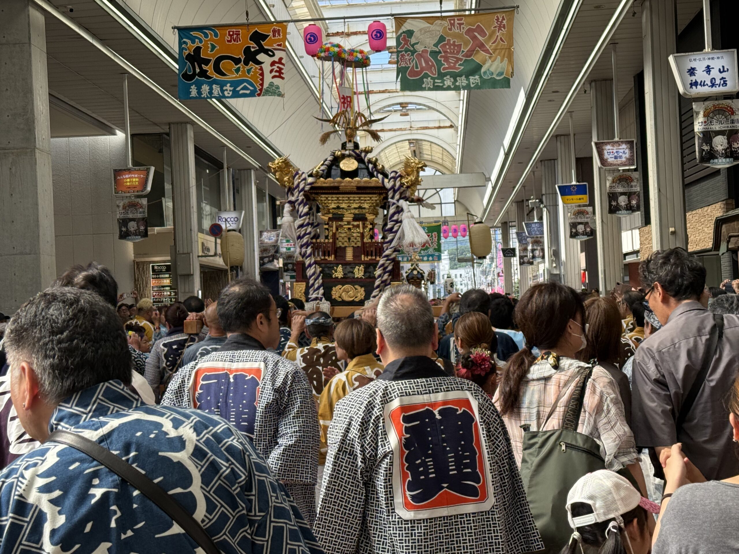 サンモール一番街のアーケード内で、背中に紋様が入った半纏(法被)を着た大勢の担ぎ手や観客が、豪華な金色の神輿を取り囲みながら渡御する熱気あふれる様子