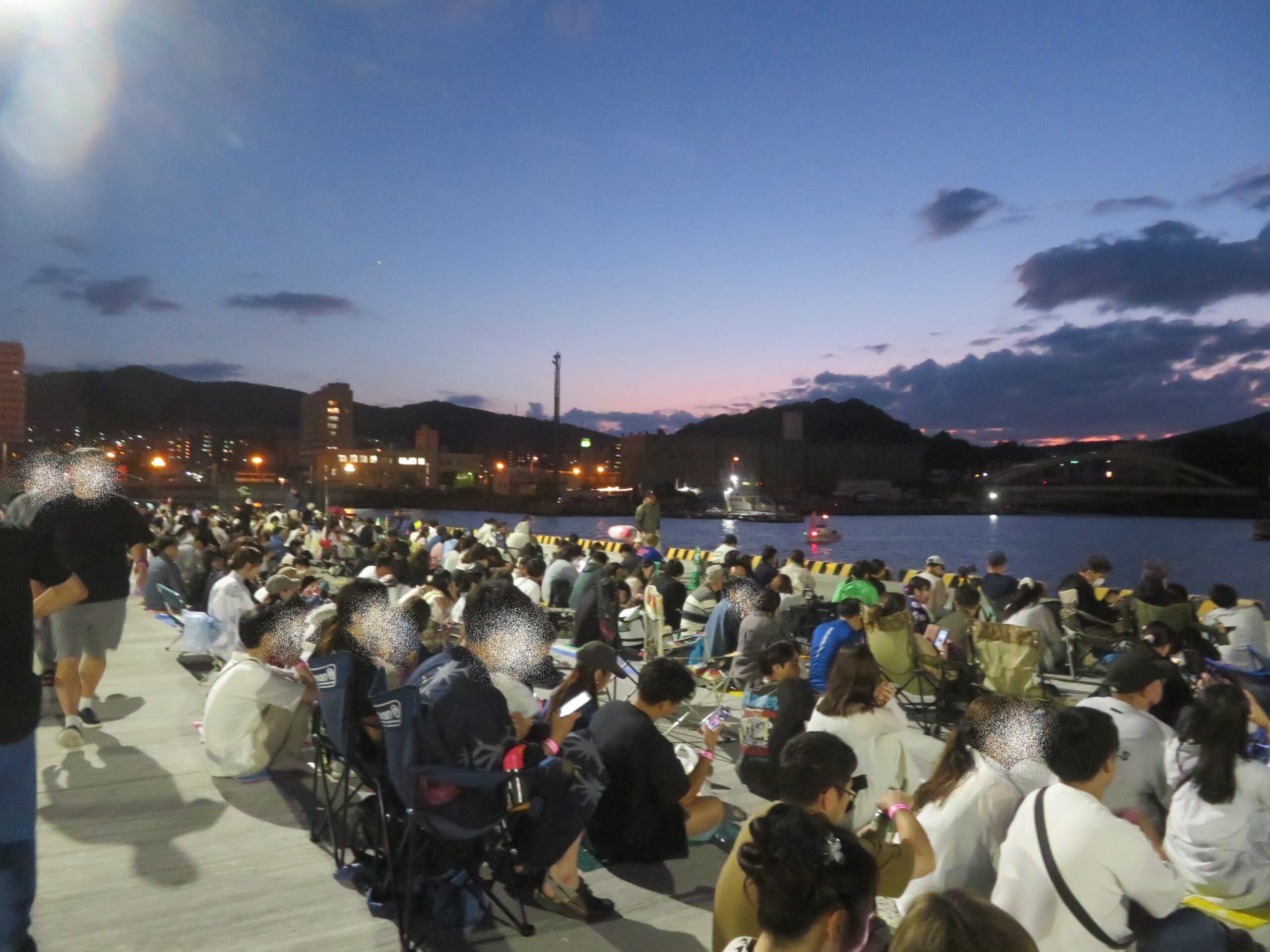 夕焼け空が広がる港の有料観覧エリアの埠頭で、アウトドアチェアや敷物に座って花火大会の開始を心待ちにする大勢の観客の様子