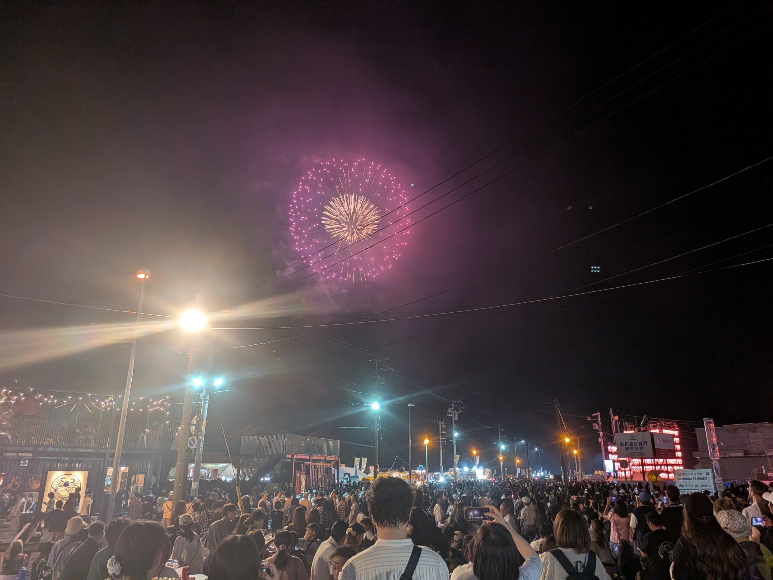 街灯や出店の明かりが輝くおたる潮まつり会場と、大勢の観客の頭上に広がる夜空に、鮮やかなピンク色の大輪花火が打ち上がる瞬間