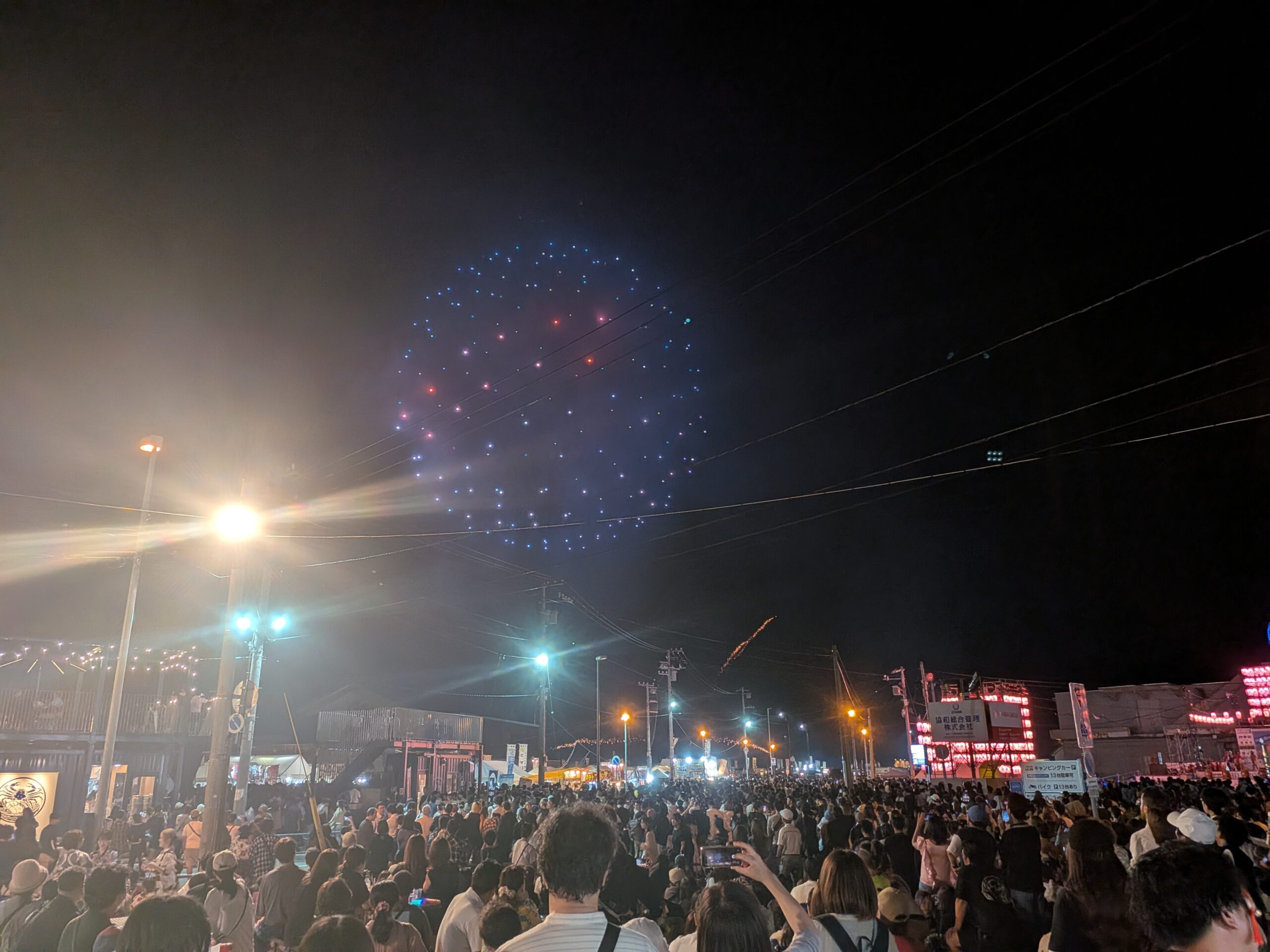 多くの来場者で埋め尽くされたおたる潮まつり会場と、街灯が輝く夜空に、青やピンクの光粒が丸く美しく広がる花火の瞬間