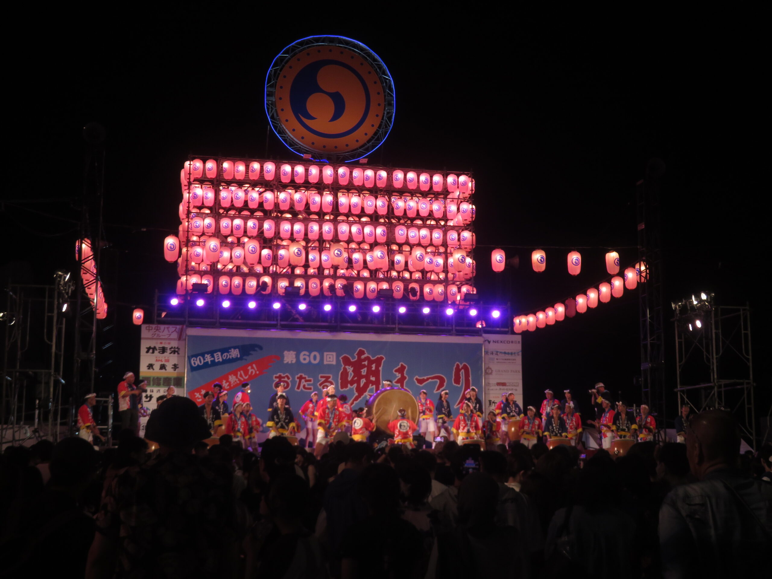 夜空にピンクの提灯とロゴシンボルが鮮やかに浮かび上がるメインステージで、閉祭式やさよなら潮音頭に続いて大勢のメンバーが一斉に太鼓を打ち鳴らし、祭りの最後を飾る様子