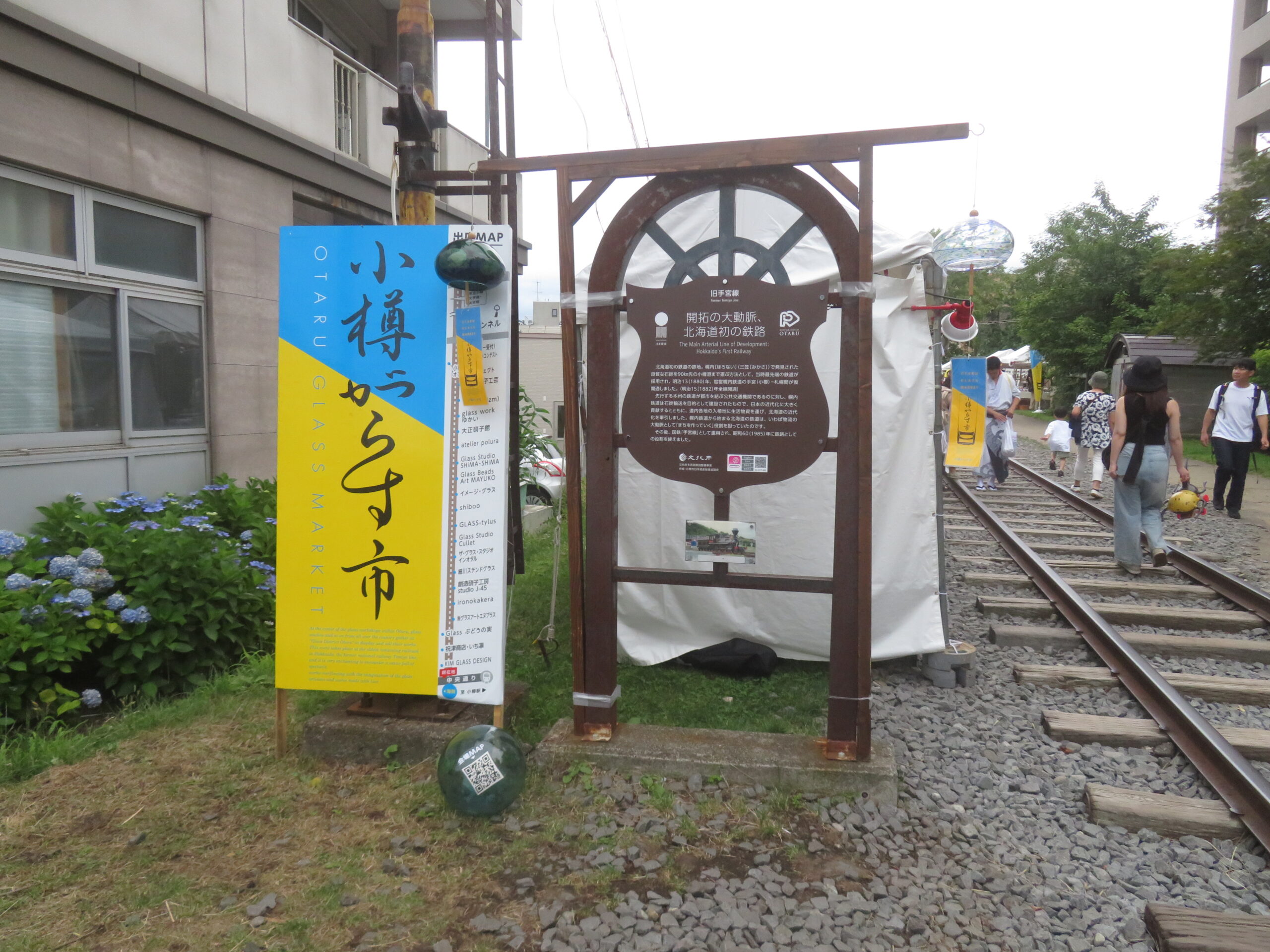 旧手宮線跡の線路沿いに設置された「小樽がらす市」の鮮やかな黄色い看板と、旧手宮線の歴史案内板。線路の上を散策する来場者の様子