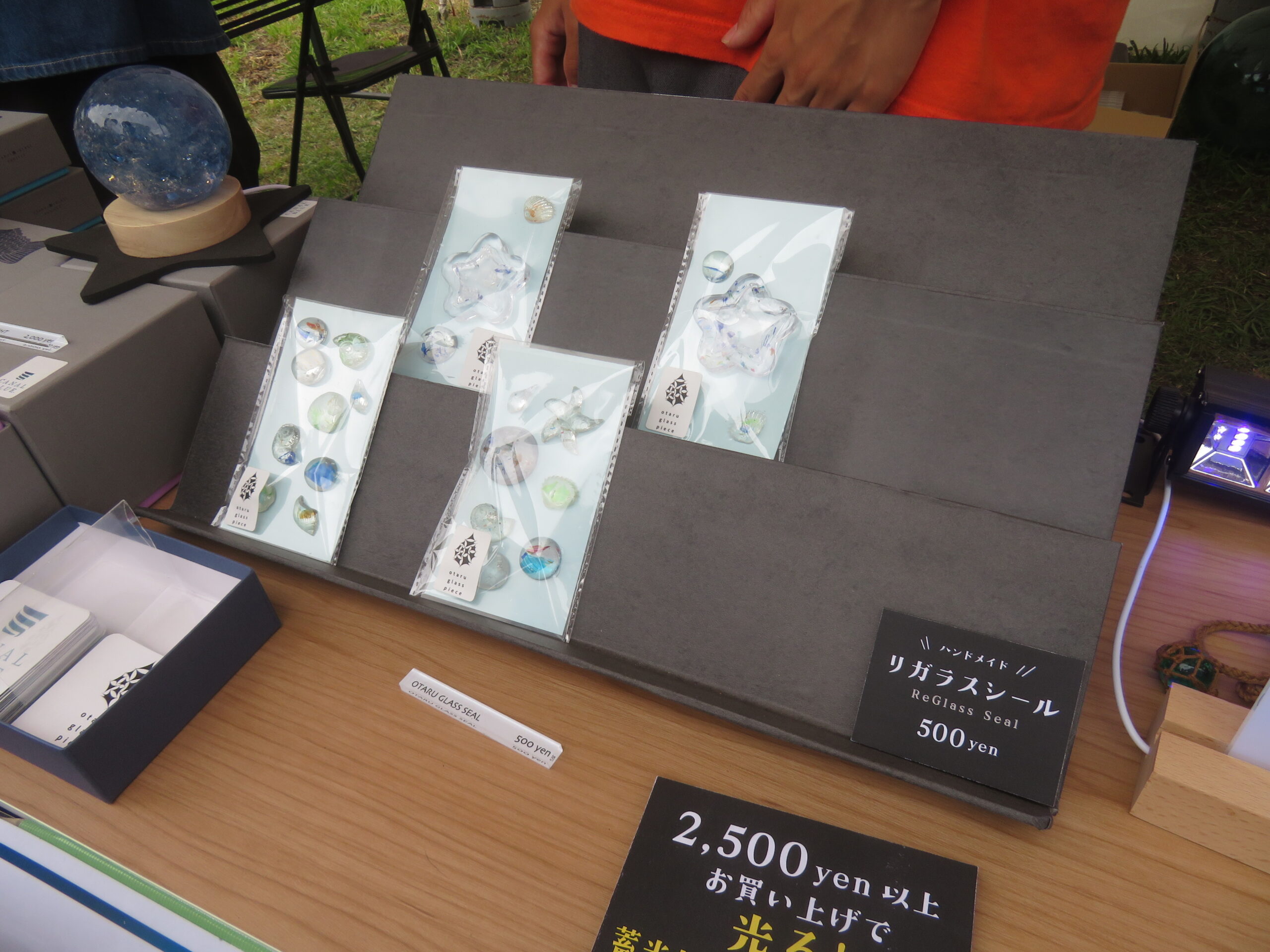 グレーのディスプレイ台に並べられた、星形や丸型のガラスパーツが入ったハンドメイドの「リガラスシール(500円)」とプライスカード