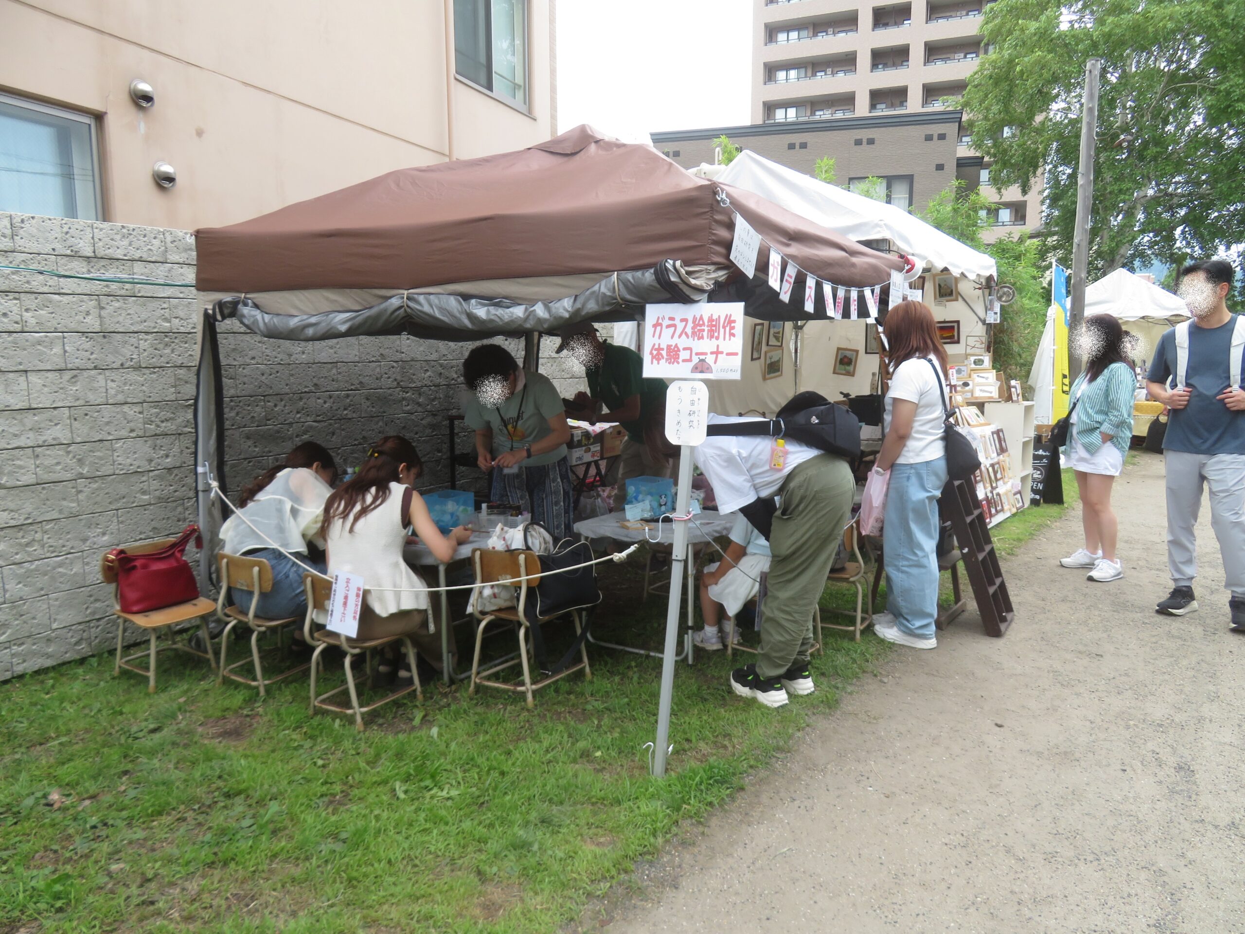 旧手宮線の野外会場で、茶色のテントの下に設置された「ガラス絵制作体験コーナー」にて、机に向かってワークショップを楽しむ参加者たちと周辺の様子