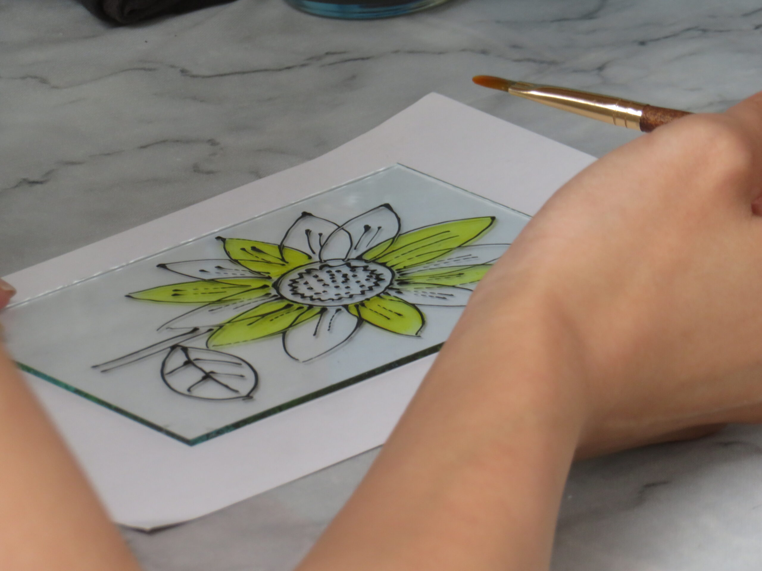 白い下絵の上に置かれたガラス板に、黒い線で描かれた花の図案。手に持った筆で、その一部に黄色い絵の具を塗り、ガラス絵を制作するワークショップの様子