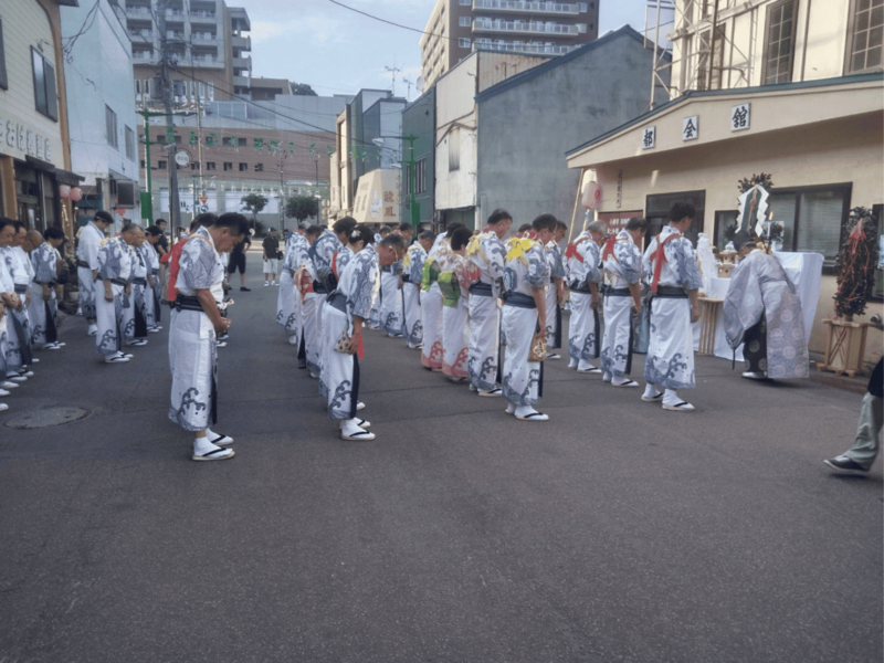 梁川商店街振興組合前に設置された祭壇に向かい、浴衣姿の参加者たちが頭を下げて潮ふれこみの安全祈願祭を行う様子