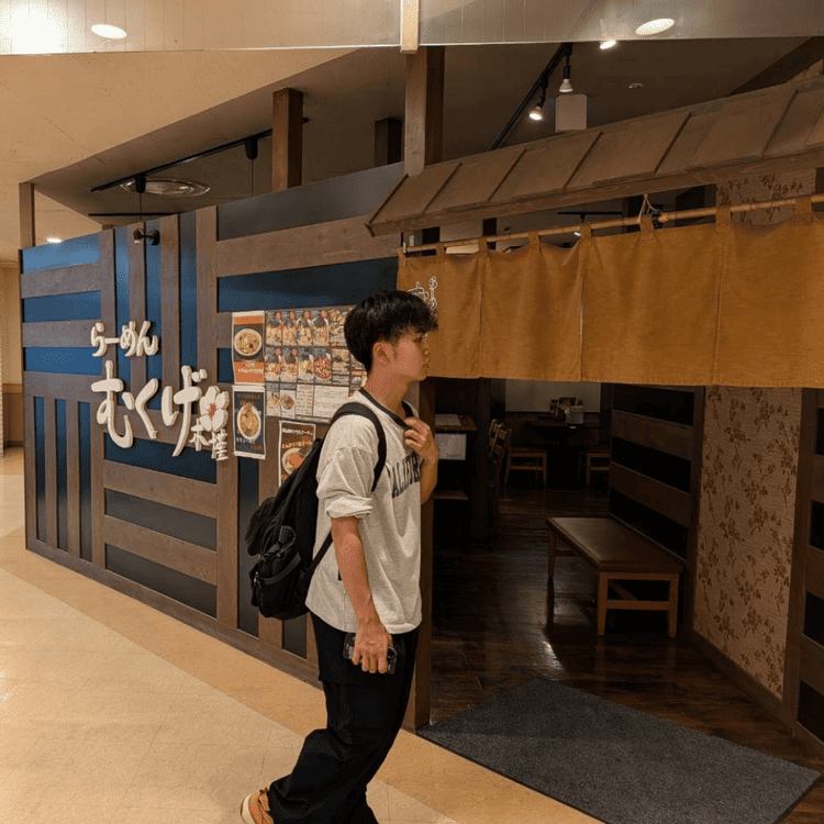 「長崎屋小樽店」内にある「らーめん むくげ」の店舗前と、店に入ろうとするリュックを背負った大学生の姿